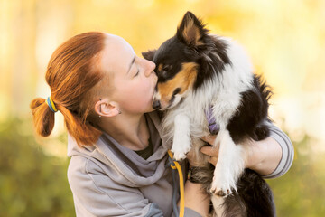 A woman with red hair hugging a shetland sheepdog outdoors. Dog with owner on Autumn blurred yellow...