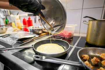 A close-up of a chef pouring batter into a hot pan, with blurred kitchen utensils and ingredients in the background. Perfect for culinary, cooking, and food preparation themes.