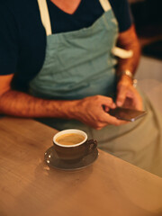 A person in an apron enjoys coffee while using a smartphone at a cozy café in the morning light