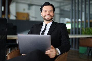 A man in a suit and tie is sitting in a chair with a laptop in front of him. He is smiling and he...