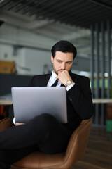 A man in a suit is sitting in a chair with a laptop open in front of him. He is deep in thought, possibly working on a project or problem. Concept of focus and concentration