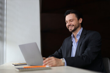 A man is sitting at a desk with a laptop open in front of him. He is smiling and he is enjoying himself