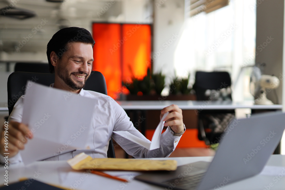 Poster a man is sitting at a desk with a laptop and a stack of papers. he is smiling and he is happy