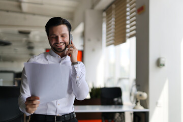 A man is talking on his cell phone while holding a piece of paper. He is smiling and he is in a...
