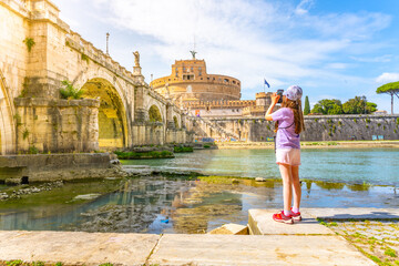 A young girl captures a photo of Castel SantAngelo and a bridge in Rome with her smartphone on a bright sunny day. Rome, Italy