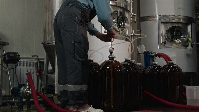 Caucasian male worker wearing overalls and white clogs standing by large stainless steel tanks while working with several dark-colored glass carboys with red hoses connected to them