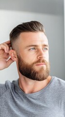 Obraz premium A man with a well-groomed beard and stylish haircut, applying pomade to his hair in front of a mirror, highlighting modern male grooming