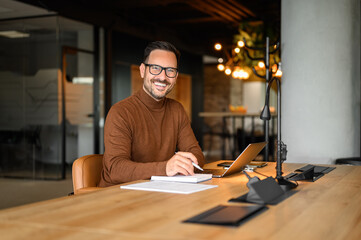 Portrait of smiling male accountant with laptop and note pad analyzing revenue report at desk in...