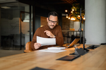 Serious male accountant in glasses examining spreadsheets and reports while working at desk in...