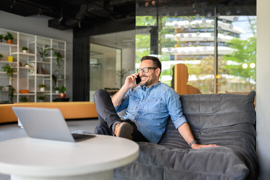 Confident sales manager discussing over phone call while sitting on comfortable sofa at his workplace