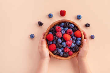 Child hands holding bowl of fresh summer berries on beige background top view. Organic and...