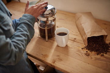 Coffee, beans and plunger with hands of woman to prepare warm beverage to drink. Strong brew, wooden table and person closeup with cup at home for fresh morning caffeine with a brown paper bag blend