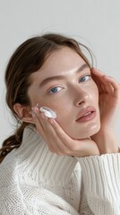 Close-up shot of woman face applying cream. Hands gently rest on her face with fingers spread apart. Model has long dark hair and wears white sweater. Plain gray background provides neutral setting.