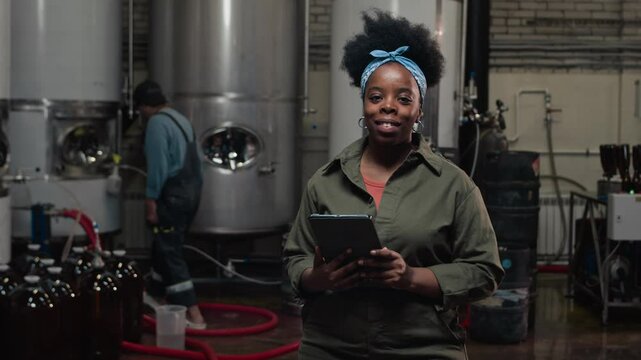 Medium slowmo portrait of smiling Black woman with digital tablet at cider production factory with Caucasian male worker and ss fermentation tanks in background