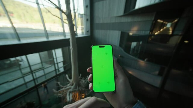 Unrecognizable Caucasian man hands holding smartphone with green screen and scrolling through social media. Guy using mobile phone on background of modern interior. Second floor of hotel hall. Indoors
