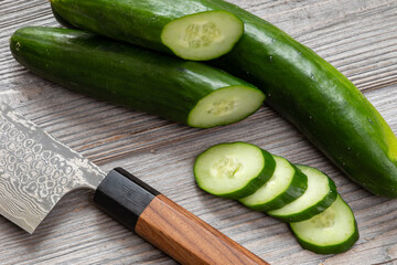 Preparing Sunomono - soy sauce pickled cucumbers. Japanese knife and fresh Japanese cucumbers, whole, halves, and slices on a rustic wooden table.