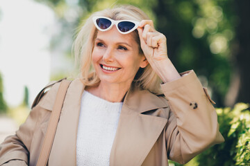 Portrait of positive happy retired woman smile spring good mood take off sunglass weekend wear beige coat outdoors outside park street
