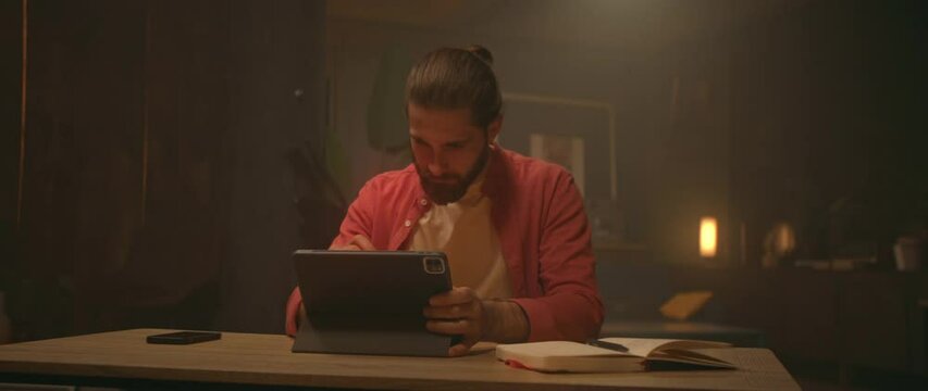 Footage young Caucasian man sitting at desk in evening and using digital tablet. Male graphic designer working in living room. Freelance work. Remote job. Camera moving around