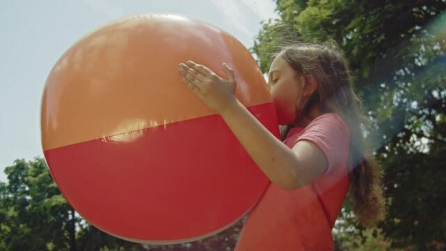 A young girl concentrates as she inflates a large beach ball in the park, enjoying a sunny day during her summer vacation. The moment captures the joy and excitement of outdoor play.