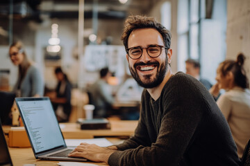 Obraz premium Smiling young man with glasses working on a laptop in a vibrant, modern office space. The atmosphere is casual yet productive, fostering creativity and collaboration.