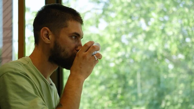 A young handsome man is sitting in a cafe and drinking coffee from a white ceramic cup. The guy is sitting at a table by the window in a coffee shop and enjoying a break alone