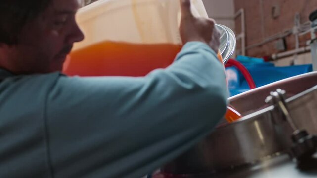 Caucasian male brewery worker wearing overalls and backward baseball cap filling large stainless steel tank with orange liquid in canister