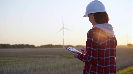 Adult woman engineer wearing white cask is taking notes on a clipboard on a field with wind turbines, as the sun sets, back view