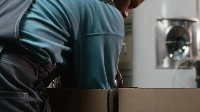 Medium shot of Caucasian male cider production plant worker placing large dark-colored glass carboy into cardboard box while preparing it for further transportation
