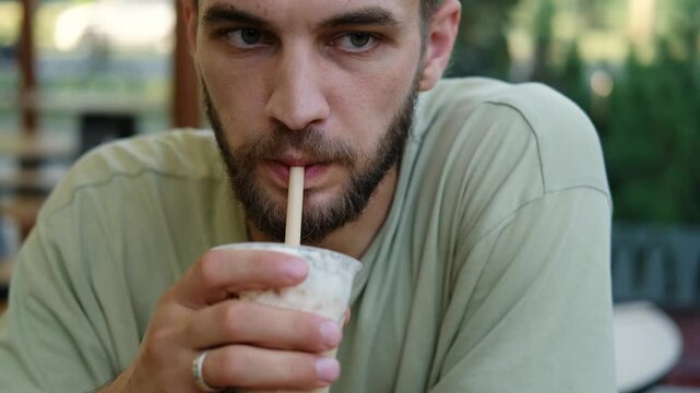 A young handsome Caucasian man is sitting in cafe terrace and drinking ice coffee or milkshake from plastic cup using a straw. Enjoying the peace of good morning