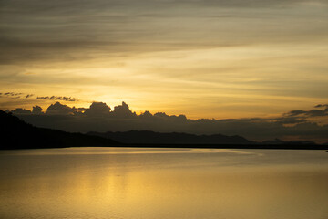 Obraz premium Golden Hour Serenity: A breathtaking view of a serene lake reflecting the golden hues of sunset, with mountains silhouetted against the dramatic sky, evokes a sense of peace and tranquility. 