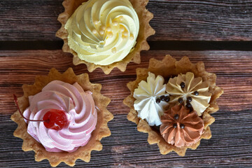 Cakes with colorful protein cream on wooden table background. Delicious tartlets with cherry, banana and chocolate flavors, shortcrust pastry. Top view.