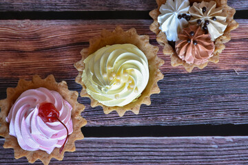 Cakes with colorful protein cream on wooden table background. Delicious tartlets with cherry, banana and chocolate flavors, shortcrust pastry. Top view.