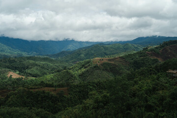 Obraz premium Emerald Peaks Under Moody Skies: A breathtaking aerial vista of lush, rolling mountains cloaked in vibrant greenery, beneath a dramatic sky of brooding clouds. 