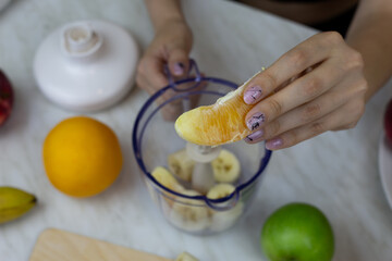 Orange slice in girl hands against background of fruits and blender on kitchen table at home, girl preparing sports cocktail with vitamins at home
