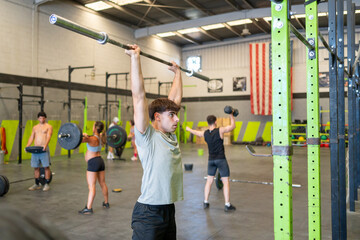 A guy warming up with a barbell in the gym before doing crossfit with a group of people training behind him.