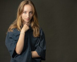 A confident young blonde woman in a loose blue shirt is looking directly at the camera with a subtle smile, posing in a professional studio with a dark grey backdrop.