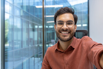 Confident young businessman smiling for selfie using smartphone in modern office environment. Displays success, positivity, and professionalism. Perfect for promoting positive workplace culture