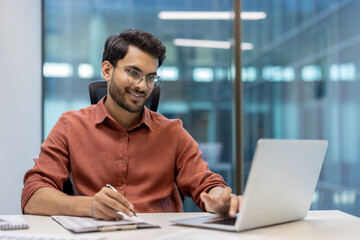 Young professional sitting at desk in modern office using laptop and taking notes. Man is focused...