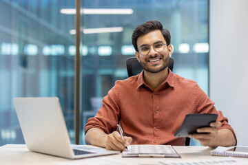 Smiling auditor in modern office holding calculator while analyzing financial data. Professional confident demeanor, laptop and phone on desk, suggesting corporate environment.