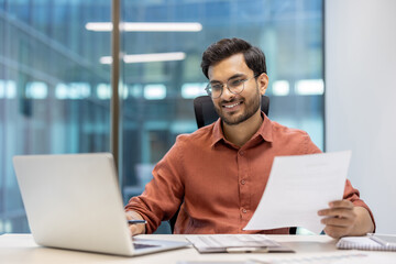 Young professional man wearing glasses working happily at laptop in modern office. Holding...
