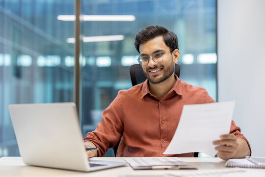 Young professional man wearing glasses working happily at laptop in modern office. Holding document, showcasing productivity and success. Captures business environment, with sense of achievement