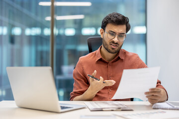 Fototapeta premium Businessman examining document while seated at office desk with laptop open. Wearing glasses and appearing concerned. Workplace setting with focus on critical thinking and decision-making.