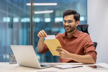 Young professional using laptop, opening envelope with joyful expression. Workplace setting with personal items, casual attire. Concept of work satisfaction, communication, modern technology.