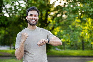 Energetic young man in park checking fitness tracker after exercise session, smiling confidently in...
