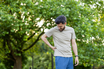 Man experiencing back pain outdoors in park with greenery; active sportsman showing discomfort while exercising on sunny day