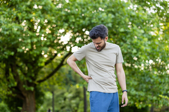 Man experiencing back pain outdoors in park with greenery; active sportsman showing discomfort while exercising on sunny day