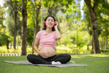 Fototapeta premium Young Asian pregnant woman enjoys listening to music with headphones in the park