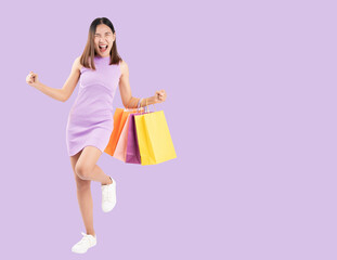 Excited young woman in a purple dress celebrates with colorful shopping bags, isolated against a purple background. This image is perfect for themes related to shopping, retail, and consumer happiness