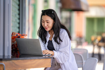 A young Asian woman in a casual white shirt works on her laptop while sitting at an outdoor café. She appears focused and engaged, enjoying the relaxed atmosphere while being productive