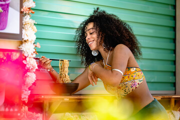 A cheerful woman of African or Afro-Latin ethnicity, wearing a vibrant outfit, enjoys eating noodles outdoors. She smiles brightly while holding a bowl, capturing a fun and relaxed moment.
